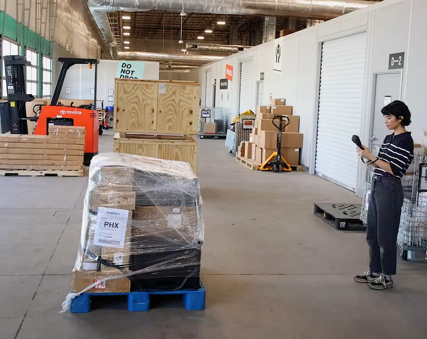 An operator holds the M2 beside a shrink-wrapped pallet on a warehouse floor, with crates and a forklift behind
